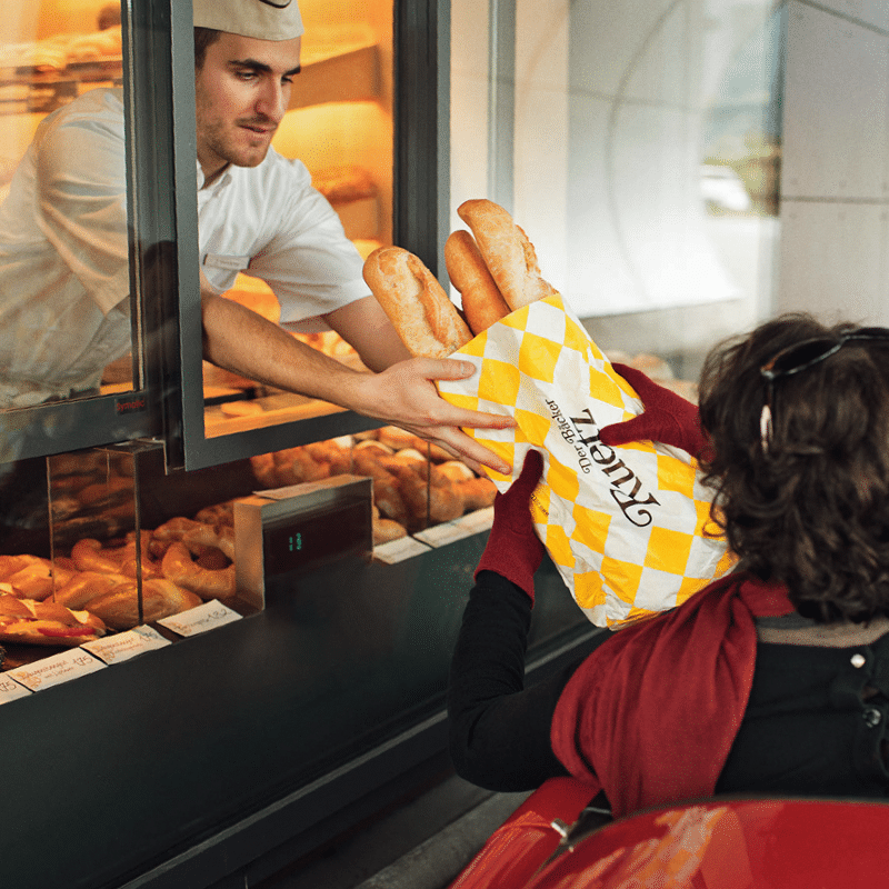 Ein Bäcker übergibt einer Frau aus dem Latenfenster eine Tüte mit Baguette für den Markenrelaunch der Bäckerei Ruetz